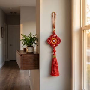 May include: A red and gold macrame wall hanging with a tassel. The decorative piece features a diamond-shaped design with a central yellow circle and white bead accents. A red tassel hangs from the bottom, and a gold cord is used for hanging.