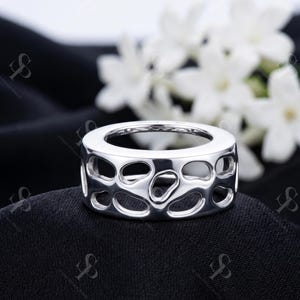 May include: A polished silver ring featuring a wide band with an openwork design consisting of irregular, rounded cutouts throughout the surface. The interior of the band is smooth and includes a small 925 hallmark stamp indicating sterling silver.