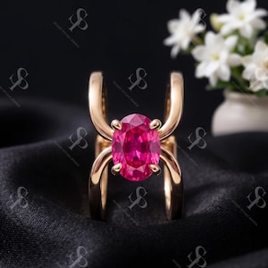 May include: A gold ring featuring a prominent oval-cut vibrant pink gemstone held by four prongs. The band consists of two parallel, curved gold segments that extend upward to secure the central stone, creating a distinct open-frame design.