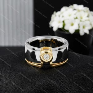 May include: A two-tone ring with a gold and silver design. The ring features a gold setting with a single, round diamond. The ring is set against a black background with a blurred floral arrangement in the background.