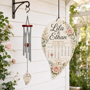 May include: A wind chime with silver metal tubes and a dark wood top, featuring a leaf-shaped wooden pendant printed with "Lila & Ethan Save the Date September 2026". Beside it hangs a large, matching leaf-shaped sign with floral rose graphics.
