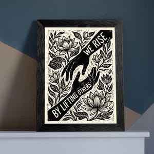 May include: A black and white framed print featuring two hands reaching toward each other amidst stylized lotus flowers and leaves. A diagonal black banner across the center contains the white text WE RISE BY LIFTING OTHERS in a bold font.