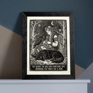 May include: A black and white framed print featuring a person with braided hair sitting in a forest with a raven and a wolf. The text at the bottom reads: SHE WEAVES THE WILD INTO SOMETHING SOFT, REMINDING THE FOREST SHE IS HOME. The frame is black.