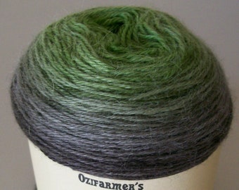 May include: A skein of yarn in a green to grey ombre colourway. The yarn is wrapped around a cardboard tube with a label that reads "Ozifarmer's Market" and a cartoon cow with the word "Rapture" below it.