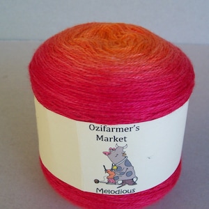 May include: A vibrant ball of yarn, featuring a colour gradient from orange to red and pink. The label displays "Ozifarmer's Market" with a cartoon cow knitting. The word "Melodious" is printed below the cow illustration.