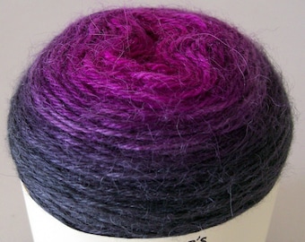 May include: A skein of yarn in a gradient of dark purple to pink with a white label that says "Ozifarmer's Market" and features a cartoon cow with a pink nose and a bell around its neck. The label also says "Rapture".