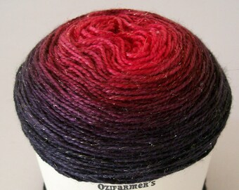 May include: A ball of yarn with a color gradient from deep red to dark purple. The yarn has a metallic sheen. The label reads "Ozifarmer's Market" and features a cartoon cow knitting. The word "Glitterata" is also on the label.