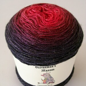 May include: A ball of yarn with a color gradient from deep red to dark purple. The yarn has a metallic sheen. The label reads "Ozifarmer's Market" and features a cartoon cow knitting. The word "Glitterata" is also on the label.