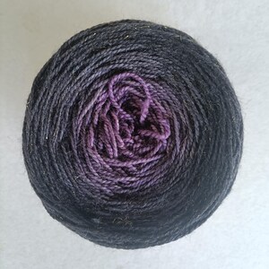Puede incluir: Una madeja de hilo con un degradado de color que va del gris oscuro al morado. El centro de la madeja es de un morado vibrante, mientras que las capas exteriores hacen la transición a un tono carbón profundo. Finos hilos dorados se intercalan en el hilo, añadiendo un brillo sutil.