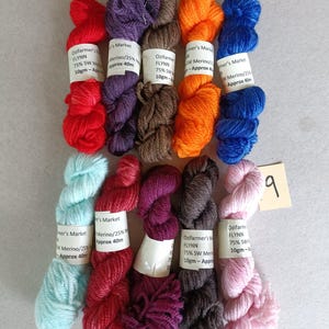 Puede incluir: Una colección de madejas de hilo de colores vibrantes, incluyendo rojo, morado, naranja, azul y rosa. Cada madeja está etiquetada con texto como "Ozifarmer's Market" y "FLYNN". El hilo está dispuesto sobre una superficie clara.