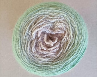 May include: A ball of yarn in a gradient of colours, shifting from a soft green to a light beige at the centre. The yarn is tightly wound, creating a textured surface, ideal for knitting or crochet projects.