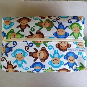 May include: Rectangular zippered pouch with a white background and a repeating pattern of cartoon monkeys in brown, blue, and green. The pouch has a beige zipper and a green fabric tab on the side. The text "Ozifamer's Market" is visible.