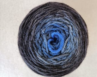 Puede incluir: Ovillo de lana con degradado de colores, pasando del azul marino oscuro a tonos de azul más claros en el centro. La lana está enrollada firmemente, creando una forma circular. La lana es probablemente de lana o fibra similar.