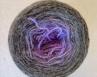 May include: A ball of yarn with a gradient colour scheme. The outer layer is a dark grey, transitioning to shades of purple and blue in the centre. The yarn appears soft and fluffy, suitable for knitting or crochet projects.