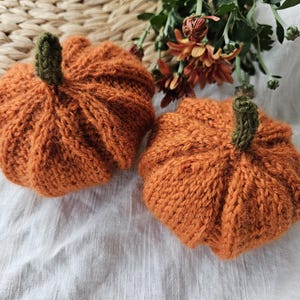 Op de afbeelding: Twee ronde, oranje gebreide pompoenen met een textuur van geribbelde oppervlakken en kleine, donkergroene gehaakte steeltjes. De pompoenen zijn gemaakt met een zichtbaar breipatroon, wat zorgt voor een zachte, ronde vorm.