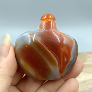 May include: A small, rounded snuff bottle carved from polished agate stone. The surface features swirling, organic bands of deep burnt orange, rust red, and translucent gray. It has a smooth, glossy finish and a matching small, rounded stopper.