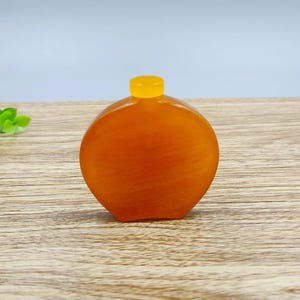 May include: A small, round, translucent amber-colored bottle with a smooth, polished finish. The bottle features a flat, circular body and a short, narrow neck topped with a bright yellow, cylindrical cap, creating a simple, uniform appearance.