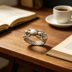 Puede incluir: Anillo plateado con un diseño detallado de rana. El anillo presenta un elemento circular central y motivos de rana a ambos lados, con una banda calada. El anillo se exhibe sobre una superficie de madera, lo que sugiere un tema de joyería o accesorios.