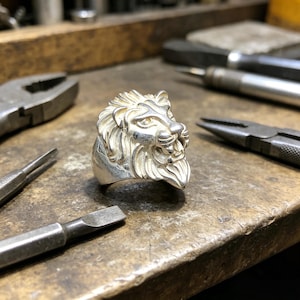 Puede incluir: Un anillo de plata con cabeza de león con detalles en la melena y rasgos faciales. El anillo está sobre una superficie de trabajo con herramientas, incluyendo alicates y destornilladores, lo que sugiere un taller de joyería. El diseño del anillo es muy detallado.