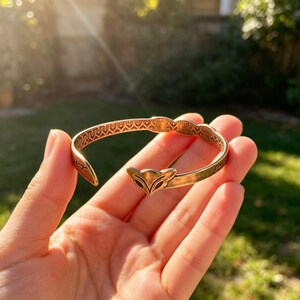 May include: A gold-toned metal cuff bracelet featuring a stylized fox head at the center. The band has an open design with an intricate geometric pattern engraved along the inner surface and visible text on the side of the metal band.