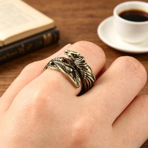 May include: A metallic, antique-gold colored wrap ring featuring a detailed design of a wolf head at one end and a textured, feather-like pattern along the band that curves around the finger, showcasing a weathered, dark-toned finish.