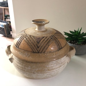 May include: A beige ceramic lidded bowl with a textured surface and geometric design on the lid. The bowl has two handles and a small, round knob on top. The bowl is likely used for serving or storage.
