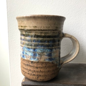 May include: Handmade ceramic mug with a unique, textured design. The mug features a tan rim and base, with bands of blue, green, and black in the middle. The mug has a curved handle and is perfect for coffee or tea.