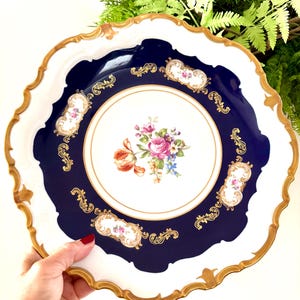 May include: A round porcelain plate featuring a white centre with a colourful floral bouquet, surrounded by a deep navy blue border. The plate has a scalloped edge trimmed in gold, with gold filigree accents and small floral cartouches.