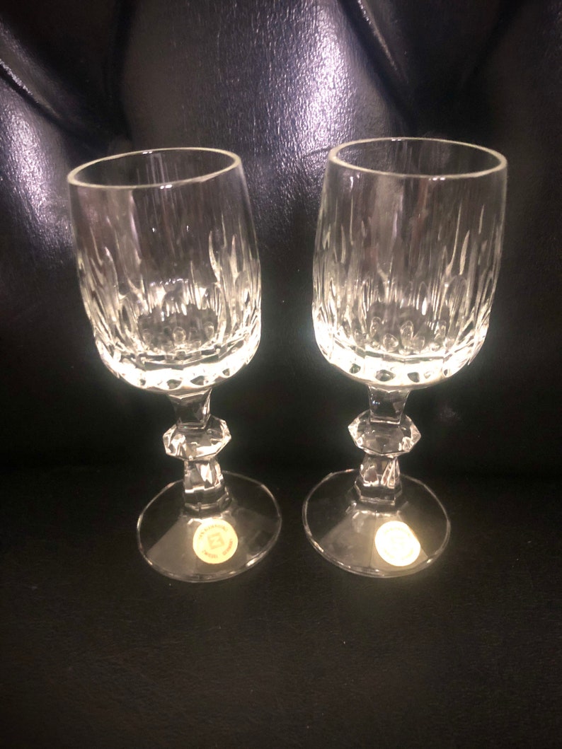 Puede incluir: Dos copas de vino de cristal transparente con cortes verticales texturizados en el cuenco y un tallo facetado. Las copas tienen una base redonda con una peque&ntilde;a pegatina. Las copas est&aacute;n sobre una superficie oscura.