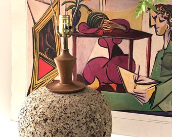 May include: A retro table lamp featuring a cork sphere base, wooden neck, and base. It has a brass lamp holder and a metal shade support. A colorful artwork is visible in the background.