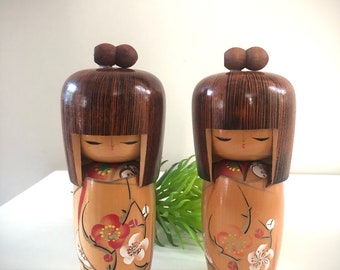 May include: Two wooden Kokeshi dolls with brown hair and a rounded top. The dolls have a light brown body with a floral design in red and white. The dolls are standing upright.