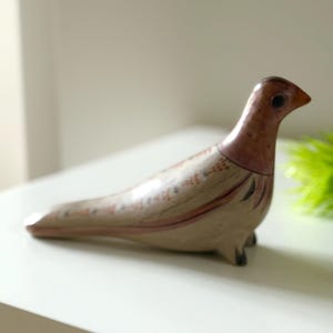 May include: A ceramic bird figurine with a long body and a slender neck. The bird is primarily beige with reddish-brown accents on the head and wings. Black markings add detail. The figurine is on a white surface.