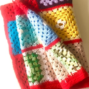 May include: A colorful, handmade crochet blanket. The blanket features a patchwork design with squares of red, blue, green, yellow, and purple. The edges are trimmed in red. The blanket is folded, showing the texture of the yarn.
