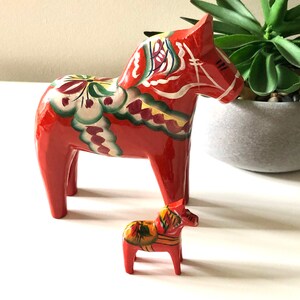 May include: Two wooden Dala horses, a traditional Swedish handicraft. The larger horse is red with floral and geometric painted details in white, green, and yellow. The smaller horse has similar details. The horses are displayed on a white surface.