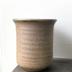 May include: A handcrafted ceramic tumbler with a textured surface and earthy tones. The cup has a wide, slightly flared rim and a cylindrical body. The exterior features horizontal striations, giving it a rustic appearance. The rim has a subtle green hue.