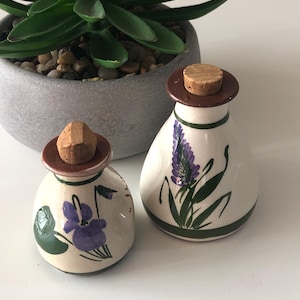 May include: Two small, white ceramic bottles with cork stoppers and brown rims. One bottle features a purple flower design, the other a lavender sprig. Both have green accents and are displayed near a potted succulent.