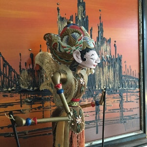 Peut inclure: Une marionnette ornée avec un visage peint, une coiffe élaborée et des vêtements traditionnels. La marionnette est placée devant une peinture abstraite aux tons orange, noir et marron. Les bras de la marionnette sont tenus par des tiges.