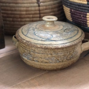 May include: A beige ceramic lidded pot with blue decorative lines and two handles. The pot is on a wooden surface, with woven baskets in the background.