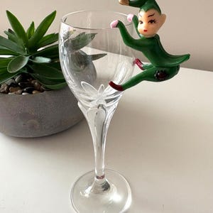 May include: A clear wine glass with a decorative green elf figurine clinging to the side. The elf has a green outfit, a gold hat, and pink accents. A succulent plant in a gray pot is in the background.