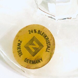 Puede incluir: Primer plano de un vaso transparente con una pegatina dorada. La pegatina dice "24% Bleikristall ZWIESEL GERMANY" con un dise&ntilde;o geom&eacute;trico. El vaso es parcialmente visible, mostrando su transparencia.