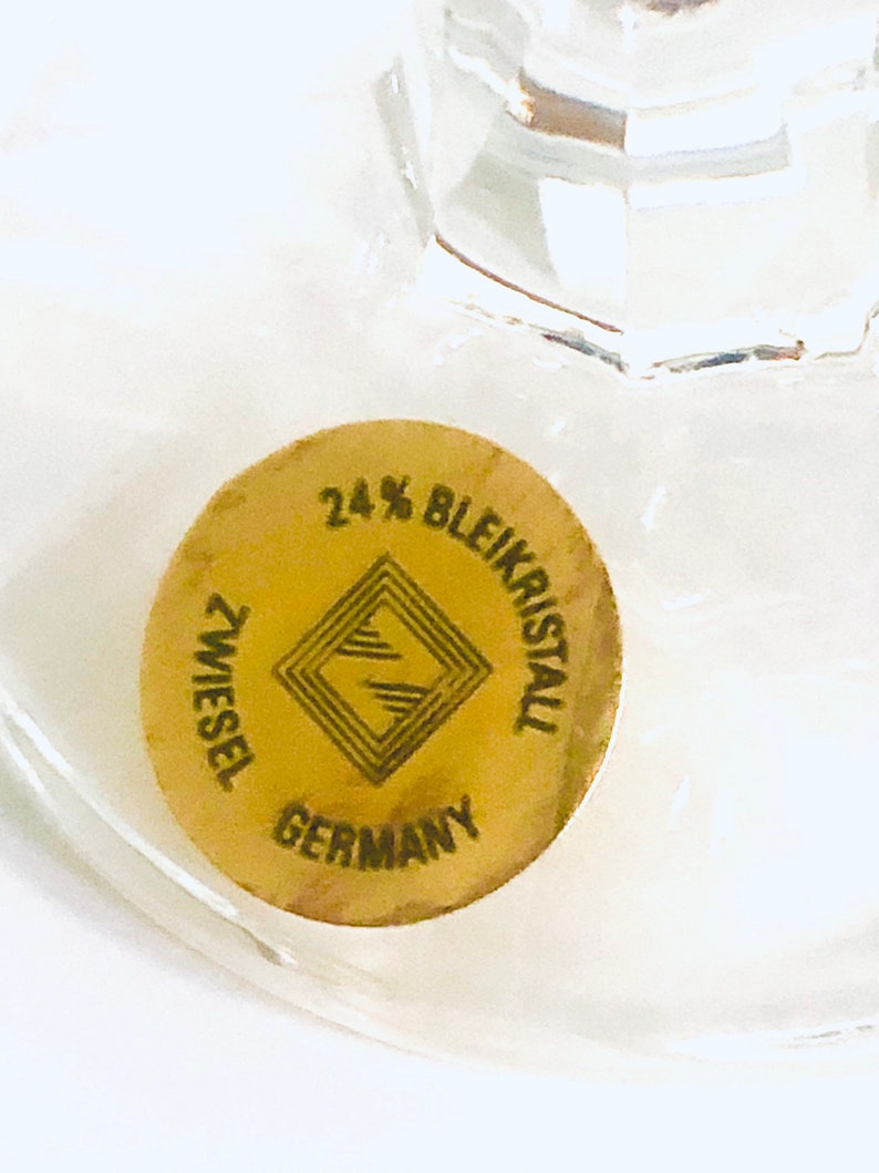 Puede incluir: Primer plano de un vaso transparente con una pegatina dorada. La pegatina dice "24% Bleikristall ZWIESEL GERMANY" con un dise&ntilde;o geom&eacute;trico. El vaso es parcialmente visible, mostrando su transparencia.