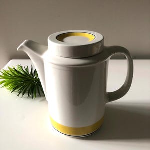 Peut inclure: Une théière en céramique blanche avec une bande jaune autour de la base et un accent jaune sur le couvercle. La théière a une poignée incurvée et un bec verseur. Un brin de feuillage vert est en arrière-plan.