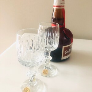 Puede incluir: Dos copas de vino de cristal transparente con dise&ntilde;os texturizados y una botella marr&oacute;n con un sello y etiqueta rojos. La botella tiene el texto "Grand Marnier" en la etiqueta. Las copas tienen pegatinas doradas en la base.