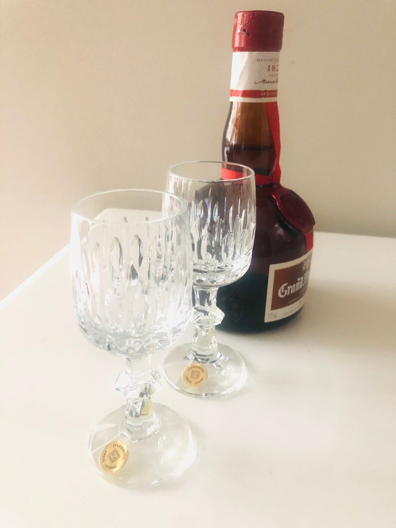 Puede incluir: Dos copas de vino de cristal transparente con dise&ntilde;os texturizados y una botella marr&oacute;n con un sello y etiqueta rojos. La botella tiene el texto "Grand Marnier" en la etiqueta. Las copas tienen pegatinas doradas en la base.