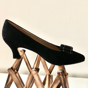 May include: A black suede pump with a low heel, featuring a decorative buckle with gold accents. The shoe is displayed on a wooden expandable rack. The heel height appears to be approximately 2 inches.