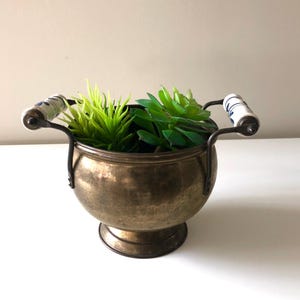Peut inclure: Un pot en laiton vintage avec un corps rond et une base en piédestal. Il comporte deux poignées en métal noir avec des poignées en céramique blanche ornées de motifs floraux bleus. Le pot est rempli de succulentes artificielles vertes.
