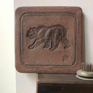 May include: A square, brown ceramic tile featuring a carved bear image. The bear is depicted walking left within a recessed border. The tile has a textured surface, suggesting it's handmade. The tile is likely made of terracotta.