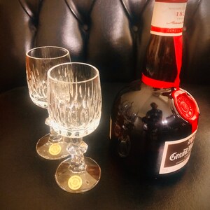 Puede incluir: Dos copas de cristal con dise&ntilde;os grabados junto a una botella marr&oacute;n oscuro de co&ntilde;ac Grand Marnier. La botella tiene un sello de cera roja y una cinta roja. El fondo es una superficie oscura y texturizada.