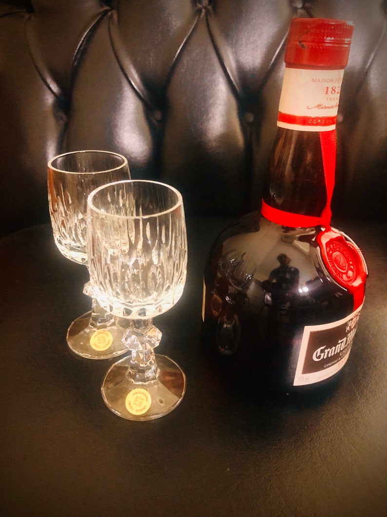 Puede incluir: Dos copas de cristal con dise&ntilde;os grabados junto a una botella marr&oacute;n oscuro de co&ntilde;ac Grand Marnier. La botella tiene un sello de cera roja y una cinta roja. El fondo es una superficie oscura y texturizada.