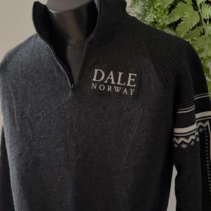 Peut inclure: Pull zippé gris foncé avec le texte "DALE NORWAY" sur un écusson rectangulaire. Le pull a une texture côtelée sur les épaules et des motifs blancs et noirs sur les manches. Le col est montant.